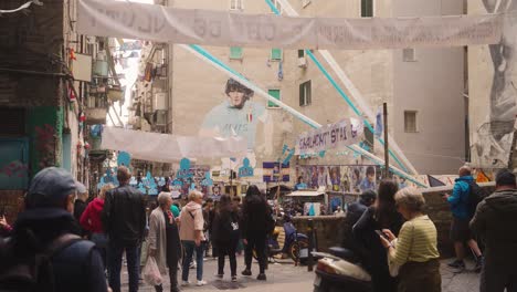 Football-fans-gather-in-memorial-square-in-narrow-street-of-Naples,-dolly-forward