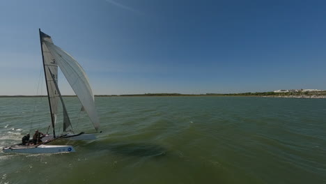 FPV-aerial-chases-catamaran-sailboat-sailing-on-green-coastal-river