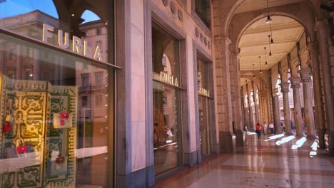 Static-shot-of-tourists-and-locals-walking-under-arches-along-sidewalk-with-rows-of-shops,-boutiques,-cafes-and-restaurants-in-Milan,-Italy-on-a-sunny-day