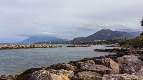 Timelapse-shot-of-rocky-seashore-with-mountain-range-in-the-background-in-Belvedere,-Italy-on-a-cloudy-day