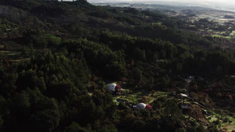 Drone-shot-of-sun-drenched-hilly-landscape-with-rolling-mist,-forest-and-small-buildings