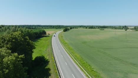 Aerial-establishing-shot-of-a-rural-landscape,-countryside-road-with-trucks-and-cars-moving,-lush-green-agricultural-crop-fields,-sunny-summer-day,-wide-drone-shot-moving-forward