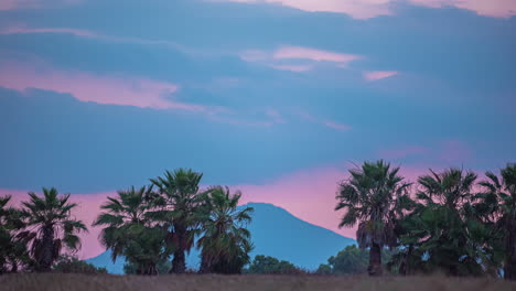 Timelapse-Unveils-the-Beauty-of-Mountain-Silhouettes-Beneath-Blushing-Skies,-with-Palm-Trees-in-the-Foreground