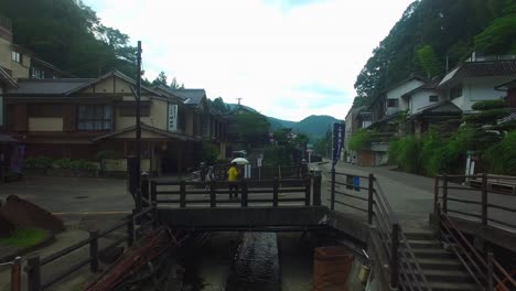 Drone-view-hovers-over-Japanese-village-watercourse,-moving-toward-a-bridge-where-two-women-pass-with-umbrellas-in-mild-rain