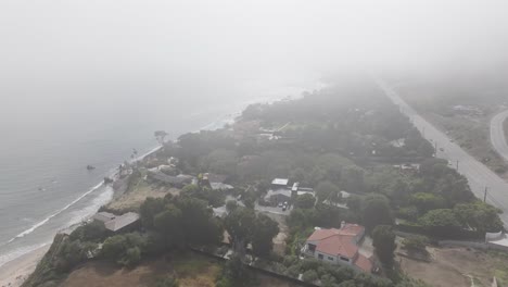Drone-cutting-through-thick-fog-or-haze-over-Pacific-Ocean-of-Malibu-neighborhood,-aerial-morning-overcast-view