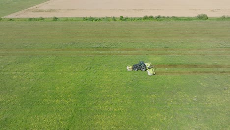 Aerial-establishing-view-of-a-tractor-mowing-a-fresh-green-grass-field,-a-farmer-in-a-modern-tractor-preparing-food-for-farm-animals,-sunny-summer-day,-wide-birdseye-drone-dolly-shot-moving-left