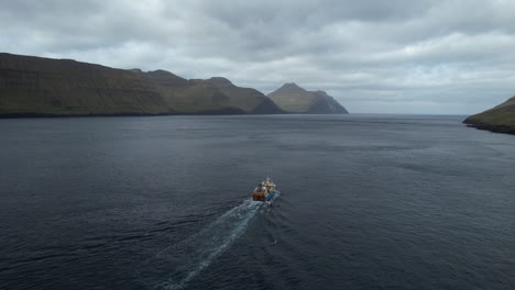Luftaufnahme-Mit-Verfolgung-Vom-Heck-Eines-Fischerbootes,-Das-Durch-Einen-Fjord-Auf-Den-Färöer-Inseln-Fährt-Und-Von-Dem-Aus-Man-Die-Großen-Berge-Sehen-Kann