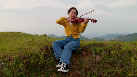 woman-peacefully-plays-the-violin-outside-while-perched-on-an-embankment,-hair-blowing-in-the-wind