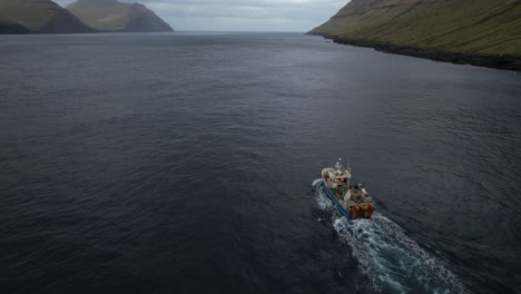 Luftaufnahme-Aus-Nächster-Nähe-Und-Mit-Verfolgung-Vom-Heck-Eines-Fischerbootes,-Das-Durch-Einen-Fjord-Auf-Den-Färöer-Inseln-Fährt-Und-Von-Dem-Aus-Man-Die-Großen-Berge-Sehen-Kann