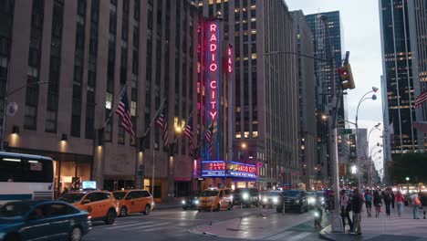 Radio-City-Music-Hall-Leuchtreklame,-Vollständige-Seitenansicht,-Mopeds-Und-Autos-Fahren-Mit-Eingeschaltetem-Licht-Vorbei