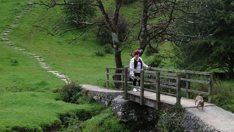 Female-with-red-dreadlocks-walking-across-rustic-wooden-bridge-crossing-stream-with-puppy,-Slow-motion