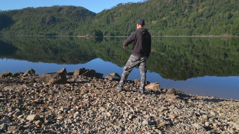 Man-stood-on-rocky-shore-of-lake-with-sharp-reflections,-surrounded-by-wooded-hills-on-sunny-summer-morning,-slow-orbit,-at-Thirlmere,-English-Lake-District,-Cumbria,-UK