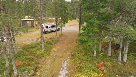 Sweden---A-White-Campervan-Tucked-Into-the-Fir-and-Birch-Forest-by-the-Lake's-Edge,-with-Breathtaking-Autumn-Colors-in-October-Under-a-Cloudy-Sky---Drone-Flying-Forward