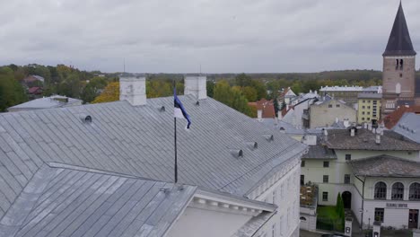 Drone-Volando-Hacia-Atrás-Lejos-Del-Edificio-Principal-De-La-Universidad-De-Tartu-Y-En-La-Parte-Superior-Hay-Una-Bandera-Estonia-Y-Al-Fondo-Podemos-Ver-El-Hermoso-Otoño-En-Tartu