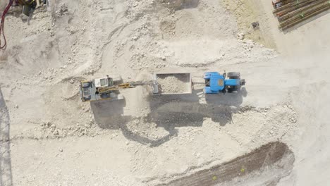 Aerial-top-down-shot-over-a-JCB-removing-soil-and-putting-it-on-a-blue-tractor-inside-a-construction-site-at-daytime