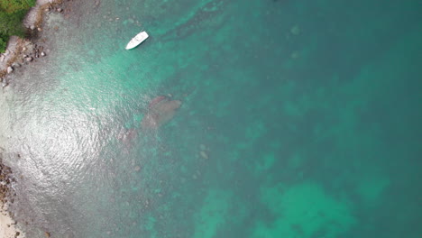 Aerial-drone-footage-with-a-top-down-view-of-turquoise-sea,-boats,-and-clear-water-revealing-the-seabed-by-a-rocky-beach