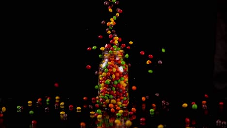 Colorful-Hard-Shelled-Candies-Are-Dropped-Into-Overflowing-Glass-Jar-on-Black-Glossy-Surface