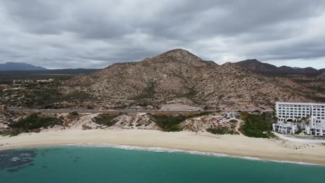 Aerial-backwards-view-of-the-beautiful-coastal-shoreline-in-los-cabos-with-a-view-of-the-sandy-beach,-a-busy-coastal-road-and-the-turquoise-sea-and-the-renowned-Marquis-hotel-during-a-mexico-vacation