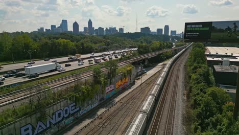 Luftaufnahme-Eines-Angehaltenen-Zuges-Auf-Der-Eisenbahn-Neben-Einer-Stark-Befahrenen-Autobahn-Und-Der-Skyline-Von-Atlanta-City-Im-Hintergrund