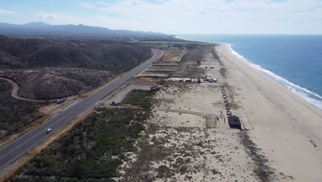 Aerial-dolly-shot-in-front-of-the-picturesque-coast-of-Cabo-San-Lucas-in-todos-santos-along-a-coastal-road-towards-Baja-California-Sur-in-Mexico-with-a-view-of-the-beach,-moving-cars-and-the-blue-sea