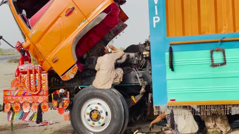 Un-Joven-Sentado-Sobre-Un-Neumático-Inspeccionando-El-Motor-De-Un-Camión-En-Pakistán
