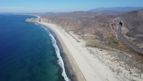 Aerial-dolly-shot-over-the-coast-of-cabo-san-lucas-of-a-pristine-beach-and-building-sea-with-calm-waves-along-a-coastal-road-on-dry-landscape-on-a-sunny-day-in-mexico