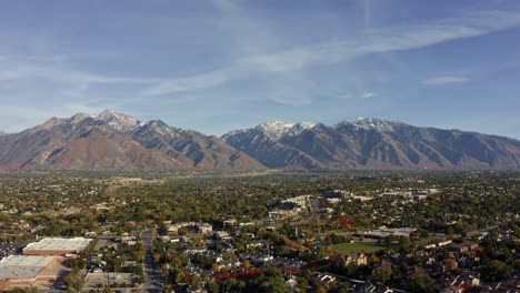 Right-trucking-tilt-down-aerial-drone-wide-landscape-shot-of-the-stunning-snowcapped-rocky-mountains-of-Utah-with-Salt-Lake-county-below-full-of-buildings-and-colorful-trees-on-a-warm-sunny-fall-day