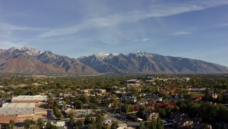 Trucking-left-aerial-drone-wide-landscape-shot-of-the-stunning-snowcapped-rocky-mountains-of-Utah-with-Salt-Lake-county-below-full-of-houses-and-colorful-trees-on-a-warm-sunny-fall-day
