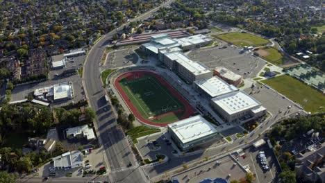 Truck-right-extreme-wide-aerial-drone-shot-of-a-large-modern-American-high-school-and-the-large-empty-track-and-football-field-next-to-a-busy-street-on-a-warm-sunny-fall-day-in-Salt-Lake-County,-Utah