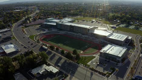 Dolly-in-tilt-down-aerial-drone-shot-of-a-large-modern-American-high-school-and-the-large-empty-track-and-football-field-next-to-a-busy-street-on-a-warm-sunny-fall-day-in-Salt-Lake-County,-Utah