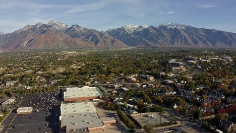 Tilt-up-right-trucking-aerial-drone-wide-landscape-shot-of-the-stunning-snowcapped-rocky-mountains-of-Utah-with-Salt-Lake-county-below-full-of-buildings-and-colorful-trees-on-a-warm-sunny-fall-day
