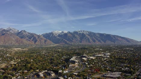 Aerial-drone-wide-landscape-shot-of-the-stunning-snowcapped-rocky-mountains-of-Utah-with-Salt-Lake-county-below-full-of-buildings-and-colorful-trees-on-a-warm-sunny-fall-day