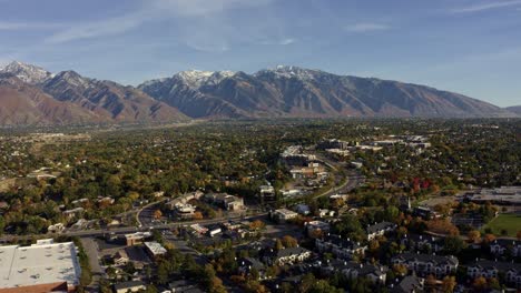 Tilt-up-aerial-drone-wide-landscape-shot-of-the-stunning-snowcapped-rocky-mountains-of-Utah-with-Salt-Lake-county-below-full-of-buildings-and-colorful-trees-on-a-warm-sunny-fall-day