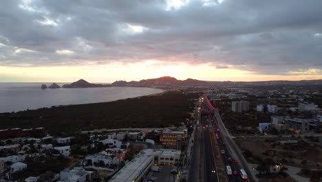 Luftaufnahme-Der-Wunderschönen-Küstenstadt-Cabo-San-Lucas-In-Mexiko-Mit-Blick-Auf-Eine-Belebte-Hauptstraße,-Einen-Sandstrand-Und-Wohnsiedlungen-Sowie-Einen-Wunderschönen-Sonnenuntergang-Zur-Goldenen-Stunde