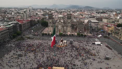 Drone-video-Mexico-City's-Zocalo,-its-iconic-cathedral,-mexican-flag-and-the-crowd-enjoying-the-square