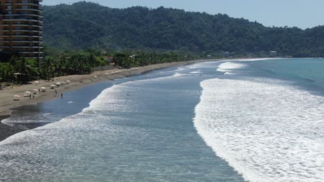 Drohnenansicht-Des-Strandes-Von-Costa-Rica,-Die-Das-Pazifische-Meer-Mit-Wellen-Und-Surfern-Zeigt,-Die-An-Einem-Sonnigen-Tag-In-Jaco-Auf-Ihren-Brettern-Sitzen