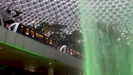 Der-Skytrain-Fährt-Neben-Dem-Fallenden-Regenwirbel-Wasserfall-Mit-Grünem-Lichtschein-Am-Flughafen-Jewel-Changi-Vorbei