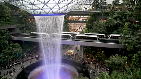 Blick-Aus-Der-Vogelperspektive-Auf-Den-Indoor-Wasserfall-„Rain-Vortex“-Am-Jewel-Changi-Airport-Singapur-Mit-Vorbeifahrendem-Skytrain