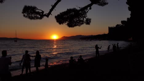 Siluetas-Oscuras-De-Personas-Y-Familias-Jugando-En-La-Playa-De-Makarska-En-Croacia-Con-Una-Hermosa-Puesta-De-Sol-Al-Fondo