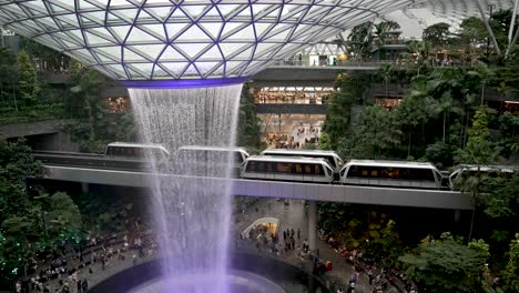 Blick-Aus-Der-Vogelperspektive-Auf-Den-Violett-Beleuchteten-Indoor-Wasserfall-Am-Jewel-Changi-Airport-Singapur-Mit-Aneinander-Vorbeifahrenden-Skytrains