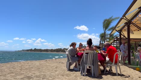 Familie-Sitzt-Und-Isst-In-Einem-Strandrestaurant-An-Einem-Sehr-Windigen-Tag-In-San-Pedro-De-Alcantara,-Sonniger-Sommertag-In-Marbella,-Spanien,-4K-Aufnahme