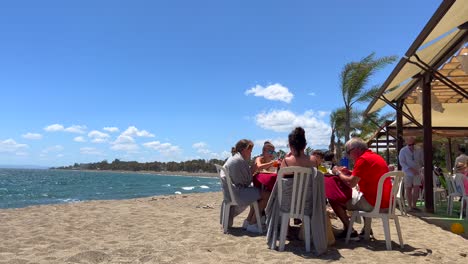 Familia-Sentada-Y-Comiendo-En-Un-Restaurante-De-Playa-En-Un-Día-Muy-Ventoso-En-San-Pedro-De-Alcántara,-Día-Soleado-De-Verano-En-Marbella-España,-Toma-De-4k