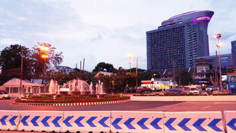 High-story-hotel-with-purple-lights-viewed-along-the-highway-roundabout-fountain-under-a-cloudy-blue-sky