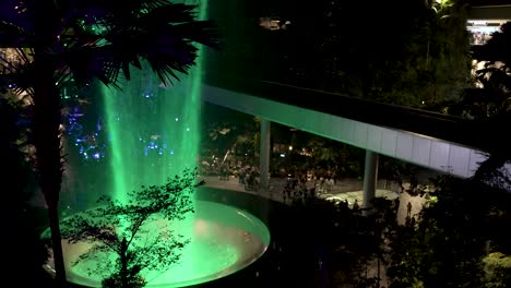 Vista-De-ángulo-Alto-Con-Vistas-A-La-Cascada-Interior-Iluminada-De-Color-Verde-En-El-Aeropuerto-Jewel-Changi-De-Singapur-Por-La-Noche