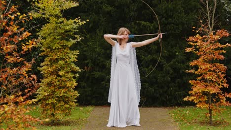 Frau,-Die-Artemis-In-Einem-Weißen-Kleid-Darstellt,-Ihren-Bogen-Zielend-Und-In-Richtung-Betrachter-Schauend