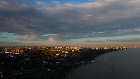 Aerial-wide-shot-over-big-river-revealing-a-city-skyline-blending-seamlessly-with-the-natural-landscape-in-sunset-warm-light,-4K50Fps