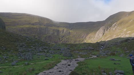 Mountain-stream-flowing-from-mountain-lake-with-man-walking-to-the-Mountains-Comeragh-Mountain-Range-Waterford-on-a-winter-day