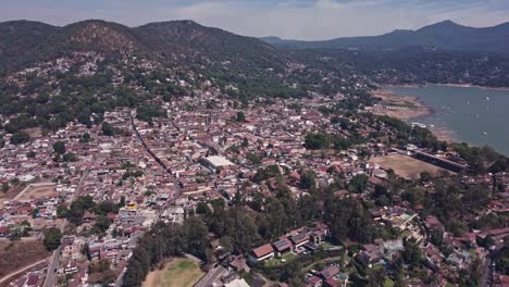 Drone-view-soaring-over-the-town-of-Valle-de-Bravo-on-a-sunny-Sunday,-capturing-the-breathtaking-sights-of-the-lake-and-the-main-church,-where-a-Ferris-wheel-spins-in-front-of-the-chapel