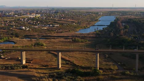 Aerial-shot-showing-multiple-cars-driving-on-a-big-viaduct-in-sunlight,-city-near-lake-in-the-background,-4k50Fps
