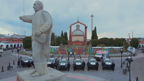 Vertikaler-Schwenk-Von-7-Polizei-Pick-ups-Und-2-Krankenwagen-Mit-Bewaffneten-Beamten-Der-Polizei,-Die-Vor-Ihren-Autos-Und-Hinter-Einer-Statue-Von-Präsident-Benito-Juárez-Aufgereiht-Sind-Und-Posieren